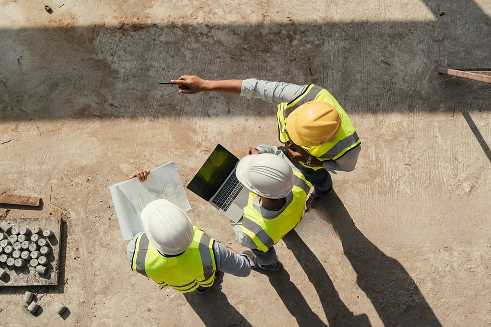 Three construction engineers in hard hats reviewing plans, laptop on site.