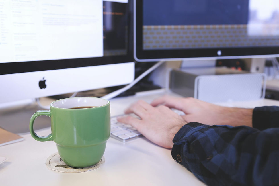 Hands typing on keyboard, green coffee mug, dual Apple monitors.