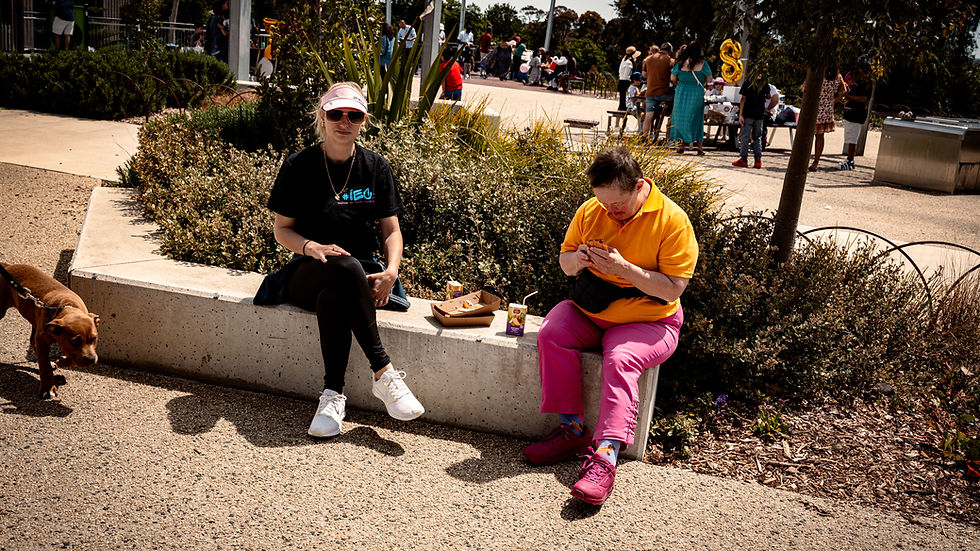 Vesna sitting outdoors beside a participant enjoying lunch during a community event, with a small dog walking nearby and other people gathered in the background.