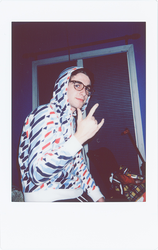 A polaroid of a Caucasian man wearing a hoodie pulled over his head and wearing glasses gives the rock and roll hand sign