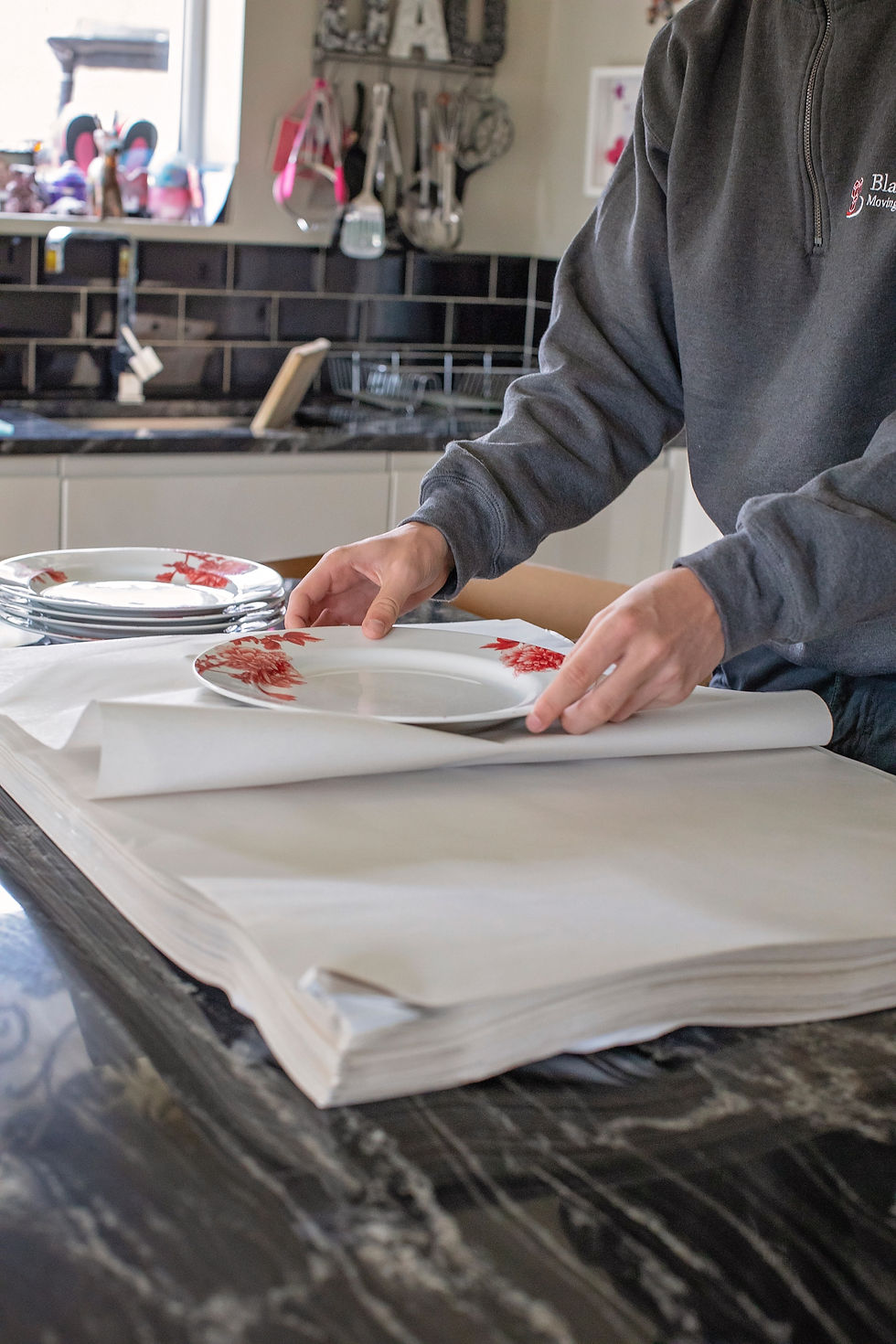 A mover carefully wrapping ceramic plates in white packing paper on a kitchen counter.