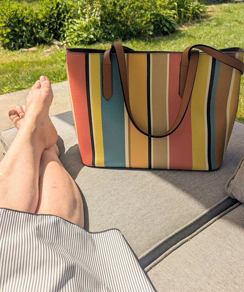 Les jambes d'une femme allongées sur un fauteuil de patio ayant à ses pieds un sac de cuir vegan aux  rayures colorées.