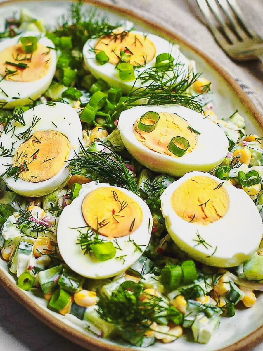 Halved boiled eggs with green onions and dill on a creamy salad with corn on a beige plate. The dish is colorful and appetizing.