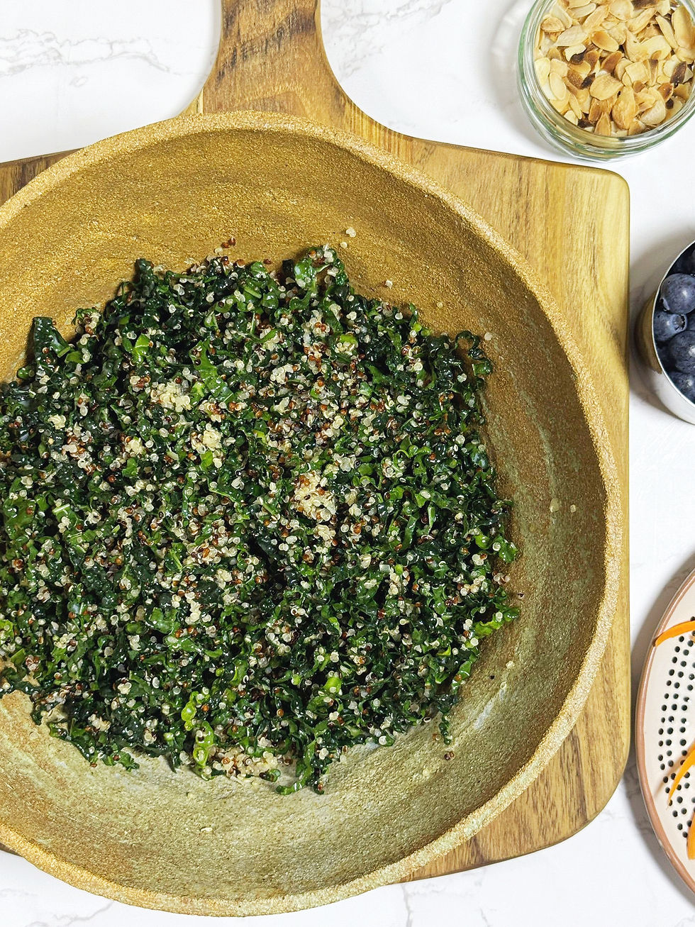 A bowl with kale and quinoa salad sits on a wooden board. Nearby are almond flakes and blueberries. Orange and black patterns on a plate.