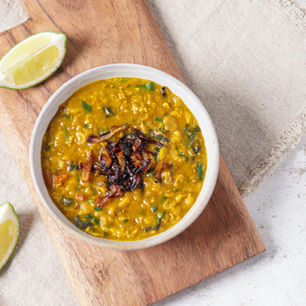 Bowl of lentil soup garnished with cilantro and onions on a wooden board, lemon wedges beside it. Rustic setting with earthy tones.