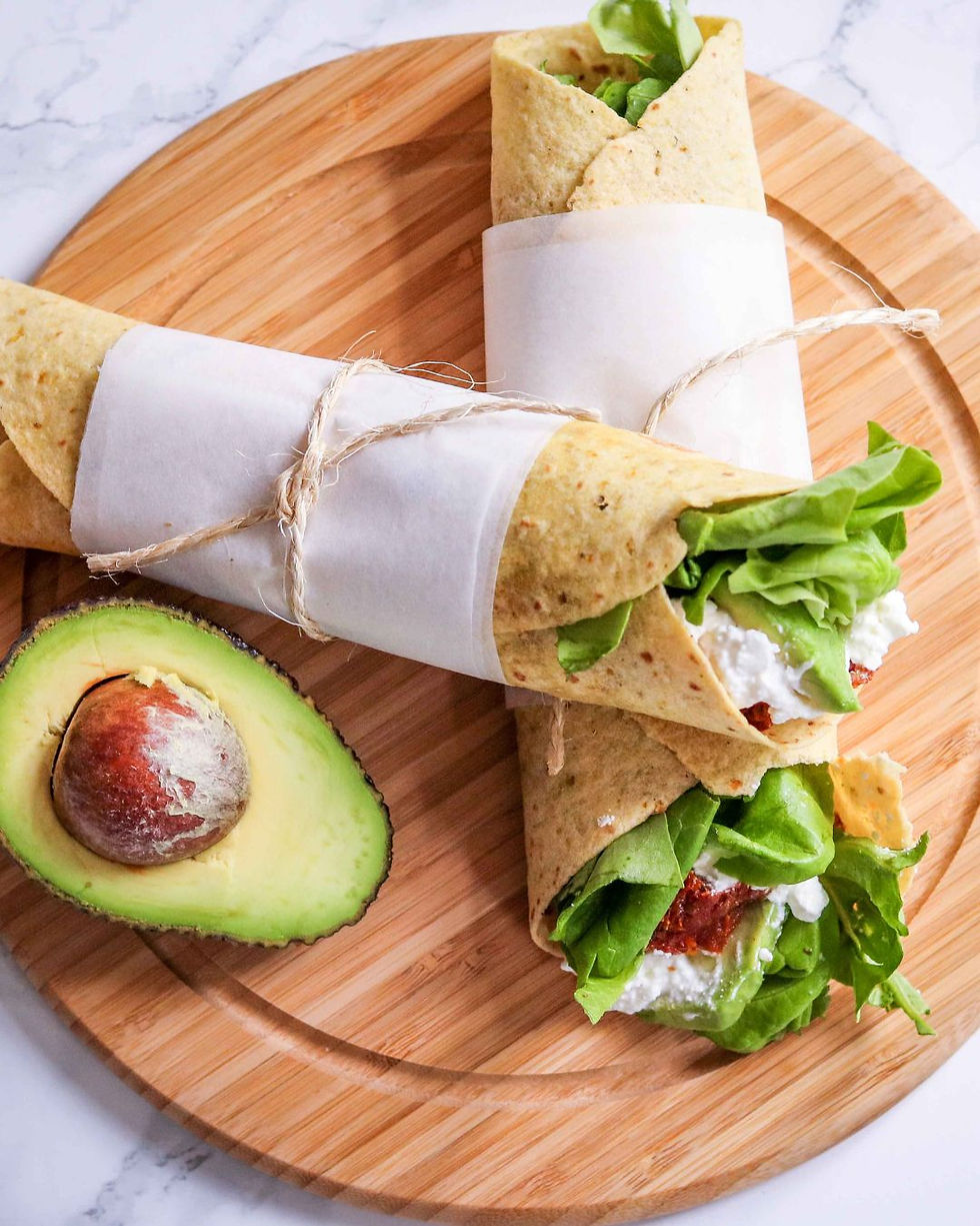 Two wraps with lettuce and cheese on a wooden board, tied with twine. Half an avocado is beside them. White marble background.