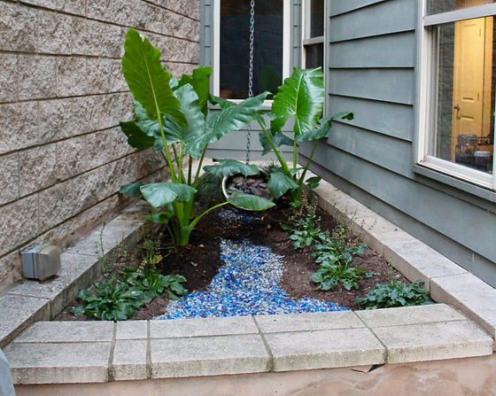 In this South Congress area yard, we removed all of the gravel in the backyard and created a whimsical design space. The door/gate and brick patio already existed but the rest of the yard was simply gravel. We scooped out all the gravel and started by spreading and tilling compost. Then, we planted shade friendly plants around an Oklahoma Patio Stone walkway which utilizes river rock from an existing feature. Lights were repaired and adjusted to highlight the new setup. In the nook by the back door, we altered the planter into a little rain garden with mixed tumbled glass. In regards to irrigation, we added Dripping Springs Ollas around the plants that need more water.