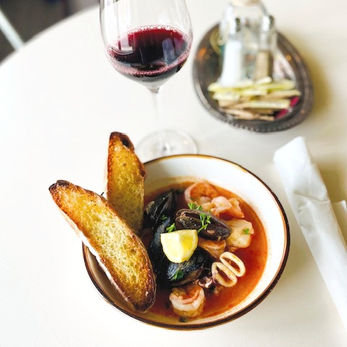 A bowl of seafood and tomato broth with two pieces of crusty bread nestled alongside the bowl.  Also pictured is a glass of red wine.