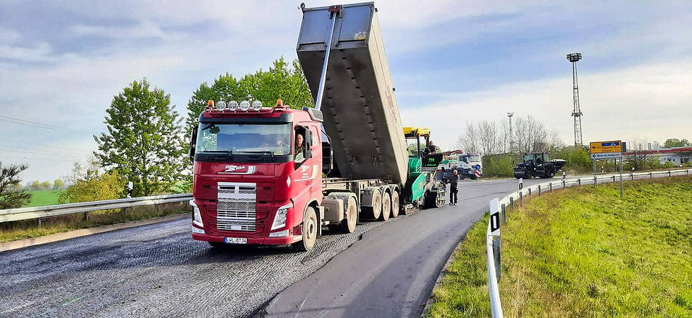 Roter LKW belädt eine grün-gelbe Asphaltiermaschine während der Aufbringung einer neuen Asphaltdecke einer Straße. Im Hintergrund steht eine schwarze Straßenwalze und ein weißer LKW, ringsum sind grüne Wiesen und Bäume zu sehen.