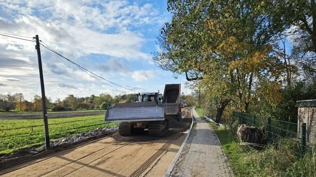 Ein Bulldozer und ein LKW bringen ein Schotterbett auf eine Straße auf, rechts ein gepflasterter Gehweg und grüne Bäume. Im Hintergrund Wiese, Wald und leicht bewölkter Himmel.