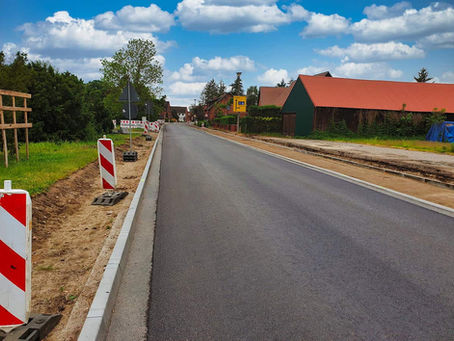 Eine Straße mit neuer Asphaltdecke und gesetzten  Bordsteinkannten, seitlich stehen rot-weiße Warnbaken, im Hintergrund Häuser mit roten Dächern und blauen Himmel mit weißen Wolken
