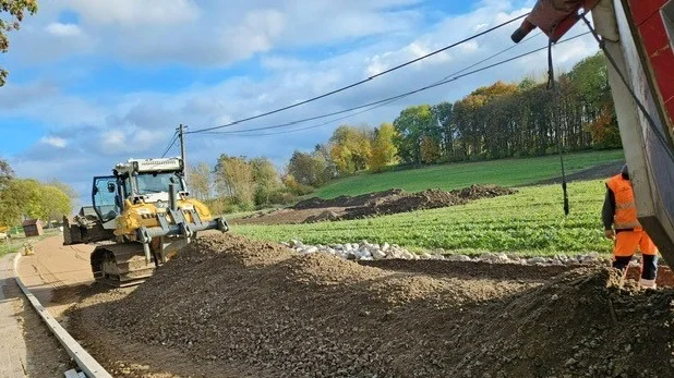 Ein gelber Bulldozer beim verteilen eines Schotterhaufens auf einer Straße. Im Hintergrund grüne Wiesen und Wälder.