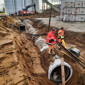 Bauarbeiter in orange Kleidung verlegen große Rohre in einem Sandgraben. Im Hintergrund ein roter Bagger und silberne Silos.