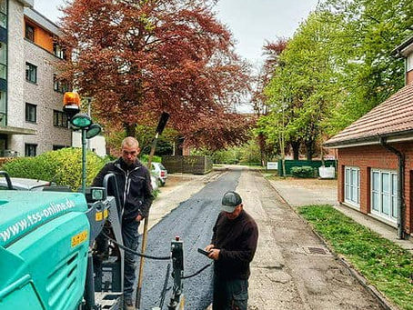 Zwei Arbeiter bedienen eine Asphaltiermaschine auf einer Wohnstraße mit Bäumen und Häusern; frische schwarze Fahrbahn.