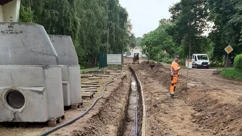 Ein ausgehobener Graben mit einer verlegten Wasserleitung, links stehen graue Betonelemente für die Kanalisation, rechts steht ein orangener Bauarbeiter. Im Hintergrund ein Minibagger umgeben von hohen Bäumen.