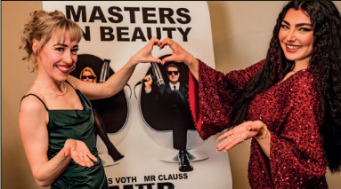 Zwei Frauen vor einem "MASTERS IN BEAUTY"-Poster formen ein Herz mit den Händen. Eine trägt ein grünes Kleid, die andere rot glitzernd.