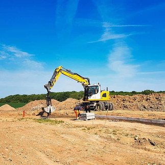 Ein gelber Bagger steht auf einem erdigen Baufeld und baggert. Im Vordergrund gesetzte Bordsteinkannten und eine Palette mit gelagerten Bordsteinkannten. Im Hintergrund Erdhäufen, Wald und blauer Himmel.