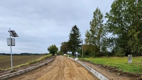 Sandbett einer Straße mit rechts und links gesetzten Bordsteinkannten. Im Hintergrund sind grüne Bäume und Wiesen unter bewölktem Himmel.