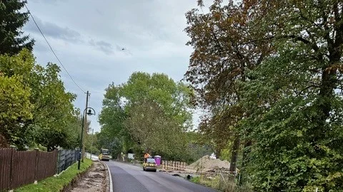 Eine neu asphaltierte Straße mit zwei Straßenwalzen im Hintergrund. Rechts einige große Bäume unter grauem Himmel.