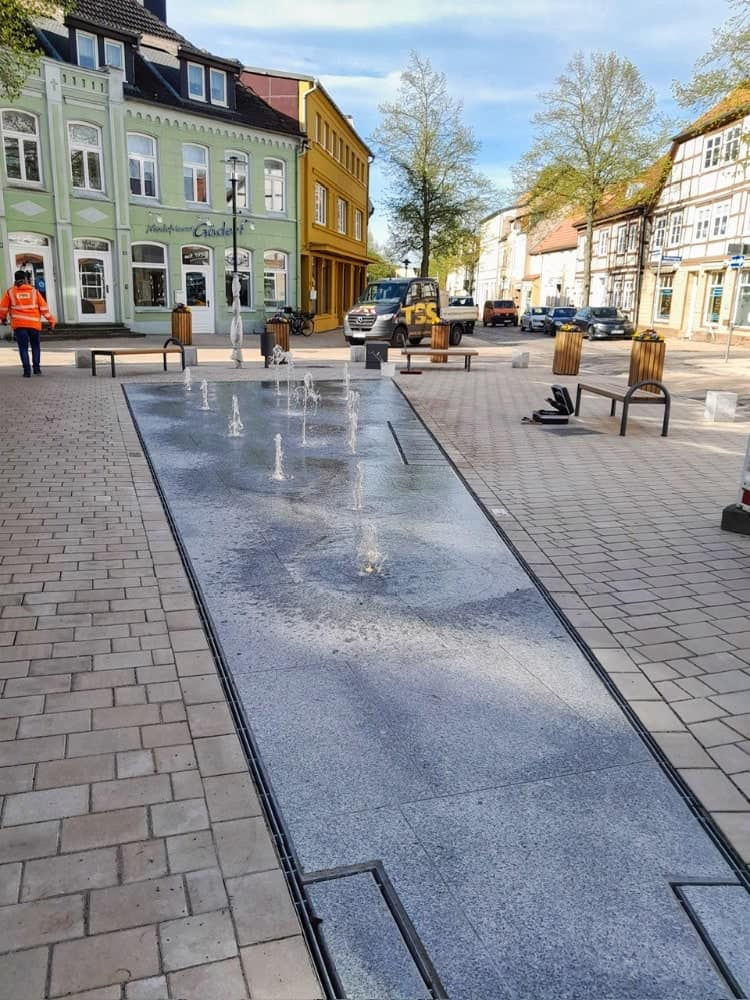 Sonniger Platz mit flachem Springbrunnen, orange gekleideter Arbeiter, pastellfarbenen Häusern und geparktem Transporter.