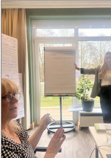 Zwei Frauen in hellem Raum bei einer Präsentation; eine zeigt auf ein Flipchart, am Fenster steht eine Pflanze.