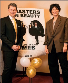 Zwei Personen in Anzügen lächeln vor einem Filmplakat "MASTERS IN BEAUTY" mit goldenen und weißen Luftballons.
