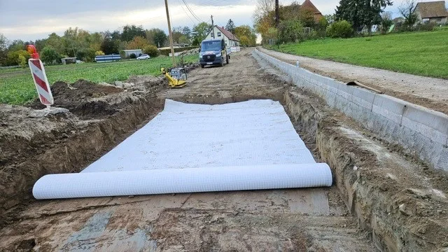 Verlegung eines Drainagefließ im Grundbett einer Straße. Daneben steht eine gelbe Rüttelplatte und im Hintergrund ein schwarzer Firmentransporter vor einer Wohnsiedlung.