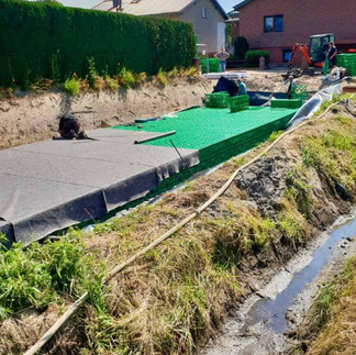 Regenwasserrückhaltungsbecken mit grünen Gitterkörben und Fließ dahinter eine grüne Hecke und ein roter Bagger