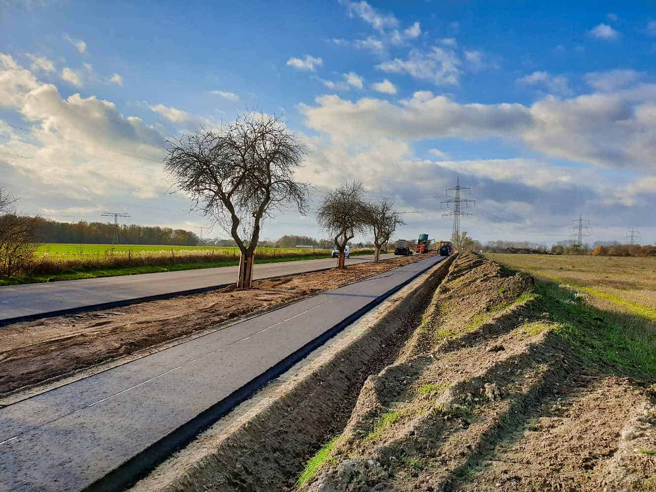 Straßenbau und Asphaltierarbeiten auf einer ländlichen Straße mit Baumreihen. Baumaschinen im Hintergrund, blauer Himmel mit vereinzelten Wolken.