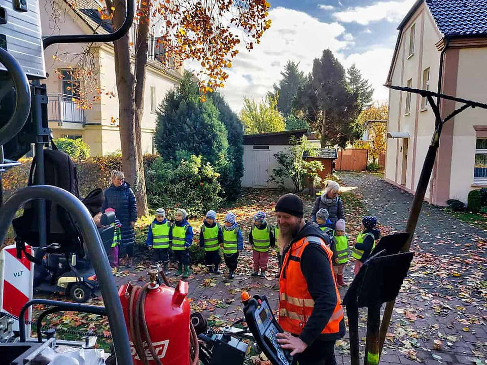 Eine Kindergartengruppe mit neon-gelben Warnwesten und zwei Erziehern beobachten einen Bauarbeiter beim bedienen einer Asphaltiermaschine. Im Hintergrund Häuser mit kleinen Büschen. 
