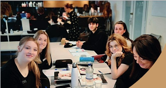 Sechs Schüler sitzen in einem Klassenraum an einem Tisch, lächeln in die Kamera; Spiegelung im Hintergrund.
