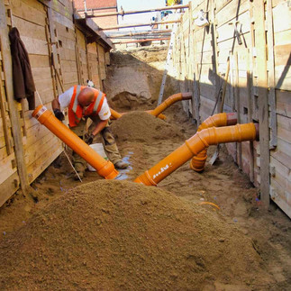 Ein Bauarbeiter mit orangener Weste und braunen Hosen steht in einem mit Holzbrettern abgestütztem Kanal und verlegt orangene Rohre