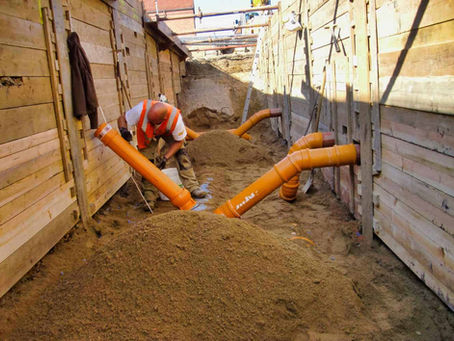 Ein Bauarbeiter mit orangener Weste und braunen Hosen steht in einem mit Holzbrettern abgestütztem Kanal und verlegt orangene Rohre