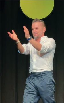 Stefan Verra im weißen Hemd und blauer Hose klatscht auf einer dunklen Bühne unter einem grünen Kreis.