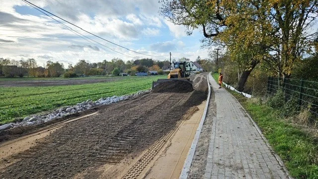 Ein gelber Bulldozer beim verteilen von Schotter auf einer Straße, rechts ein neu gepflasterter Gehweg. Im Hintergrund grüne Wiesen und Wälder.
