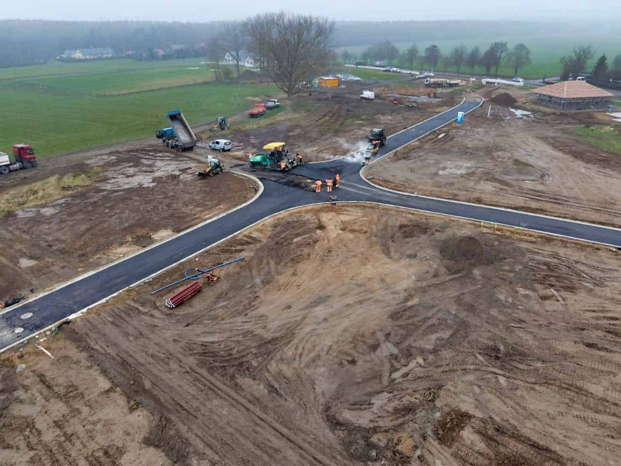 Eine erdige Grundfläche für einen neuen Wohnpark mit einer neu asphaltierten Straßenkreuzung. Mehrere LKWs und andere Baumaschinen arbeiten an der Aufbringung des neuen Straßenbelags. Im Hintergrund sind viele kahle Bäume und grüne Wiesen zu sehen.