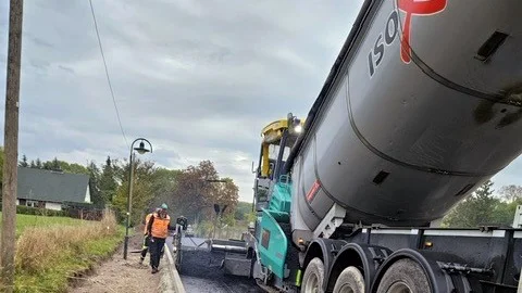 Asphaltierung einer Straße mit grüngelber Asphaltiermaschine und grauem Muldenkipper-LKW. Links daneben ein Bauarbeiter in orangener Arbeitskleidung.