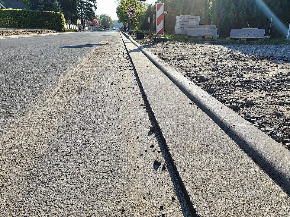 Eine Straße mit seitlich angebrachter Bordsteinkannte, im Hintergrund Paletten mit grauen Betonsteinen und einer grünen Hecke