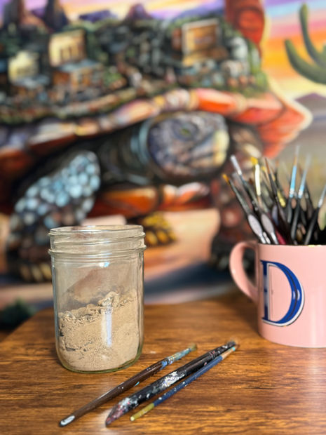 A jar of desert soil and Dani Abbott's brushes on the studio table beside Secrets of a Desert Dweller.
