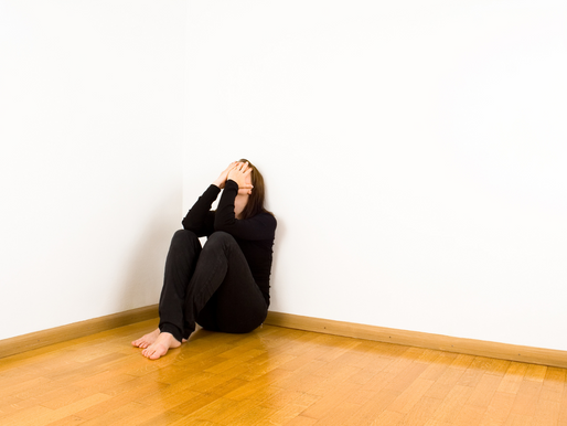 Person sits on a wooden floor in a white corner, hands covering face, wearing black clothes. Mood is distressed and isolated.