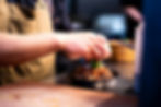 A chef's hands wearing a tan apron garnish a dark bowl of a cooked meat and pepper dish with fresh green herbs.