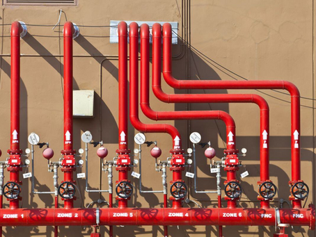 An outdoor fire protection system installed against a beige building exterior. A main horizontal red pipe along the bottom features clear white labeling for different zones, including "ZONE 1", "ZONE 2", "ZONE 5", "ZONE 4", "ZONE 6", and "FHC". Seven vertical red riser pipes extend upward, each equipped with a black control wheel, a red valve assembly, round pressure gauges, small pink spherical water motor alarm gongs, and white flow arrows marked "SPK". The five pipes on the right bend horizontally to route into the building structure.