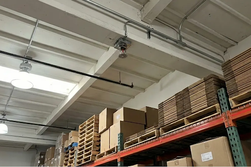 An interior view of a warehouse or storage facility with high ceilings and heavy-duty metal racking systems filled with stacked cardboard boxes, flat-folded cartons, and wooden pallets. A fire sprinkler head is mounted on a thick concrete ceiling beam, flanked by conduit pipes, black piping, and high-bay light fixtures.