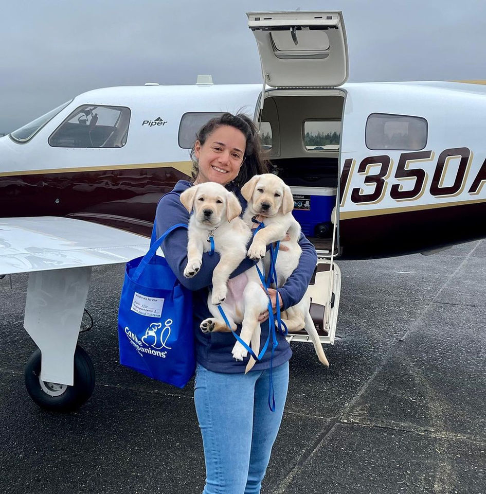 A woman poses outside of a plane holding 2 yellow Lab puppies.