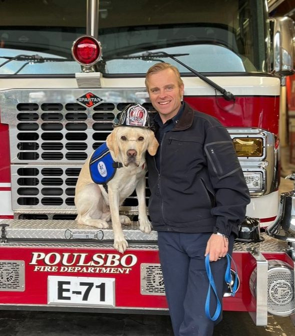 A fireman stands next to a yellow Labrador sitting on a firetruck wearing a fire hat and blue vest.