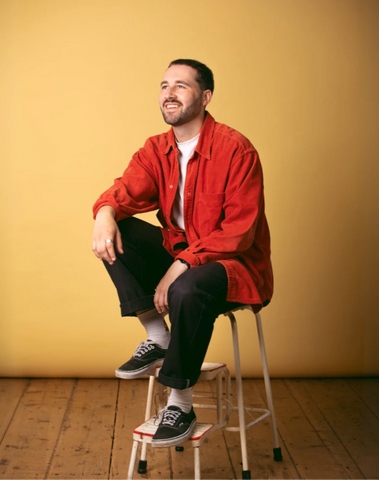 Photograph of a cool man sat smiling in front of a yellow studio backdrop