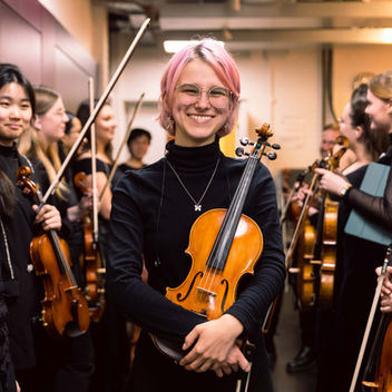 Photograph of a musician smiling and holding a violin whilst surrounded by other musicians at a music performance in Manchester