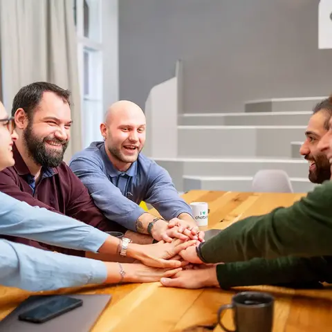 Eine Gruppe von fünf Kollegen sitzt an einem Holztisch in einem modernen Konferenzraum und legt ihre Hände übereinander, um Teamgeist und Zusammenhalt zu symbolisieren. Alle lächeln und wirken motiviert, während im Hintergrund ein schlichtes, stilvolles Interieur mit einer Checkmark-Grafik sichtbar ist. Dieses Bild ist ein inspirierendes Beispiel für Corporate Fotografie, das die Werte von Teamwork und Zusammenarbeit in einer positiven und professionellen Umgebung eindrucksvoll einfängt.