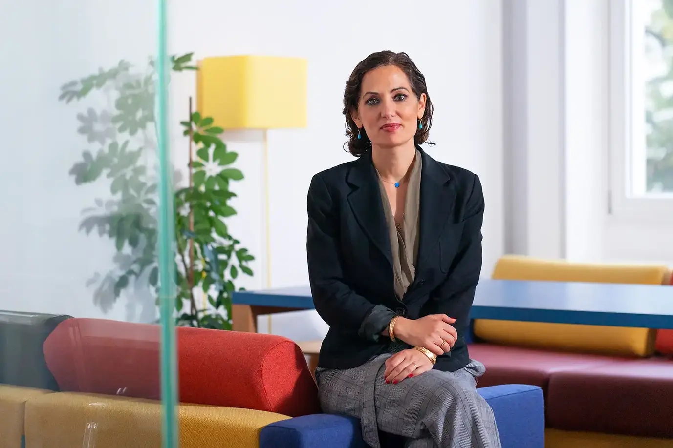 Business-Portrait einer eleganten Frau mit dunklen Haaren, aufgenommen in einer modernen, farbenfrohen Büro-Lounge. Ihr stilvolles Outfit mit schwarzem Blazer und grauer Hose unterstreicht ihre Professionalität und Kompetenz. Die entspannte, selbstbewusste Pose und das warme Ambiente machen das Bild ideal für Personal Branding und Unternehmensprofile. 