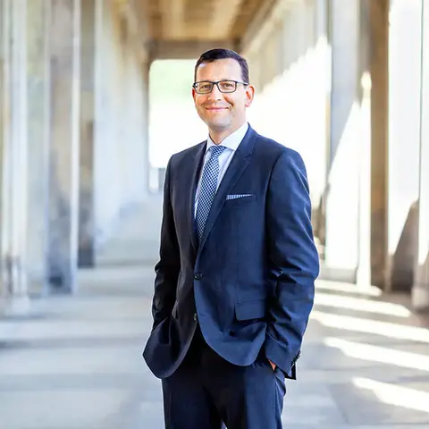 Porträt eines Mannes in einem eleganten dunkelblauen Anzug mit Krawatte, der lächelnd in einem Säulengang steht. Das natürliche Licht und die architektonischen Details im Hintergrund schaffen eine professionelle und stilvolle Atmosphäre. 